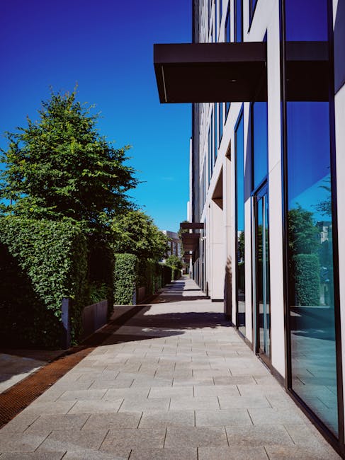 A modern outdoor garden and landscaping scene along a paved pathway beside a contemporary building with large glass windows. On the left, well-maintained green hedges and trees provide lush foliage, w